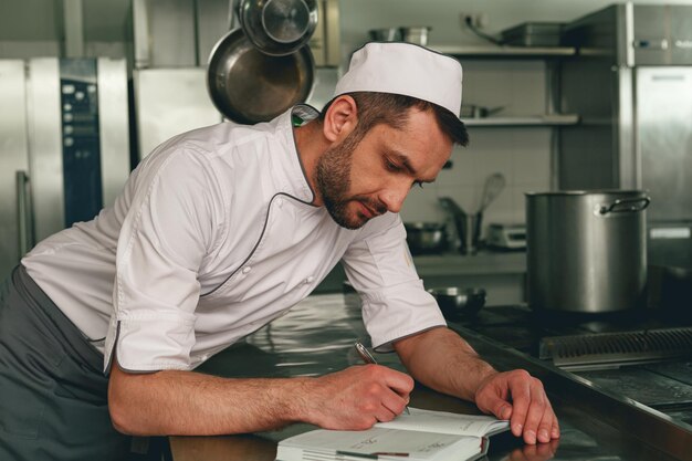 Glimlachende chef-kok in uniform die aantekeningen maakt in een notitieboekje dat op de keuken staat