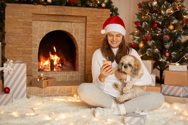 Glimlachende dame zittend op de vloer met slimme telefoon en knuffelen haar pekingese hond, dame sociaal netwerk controleren via mobiele telefoon, zit met gekruiste benen, kijkt lachend naar het scherm van het apparaat.