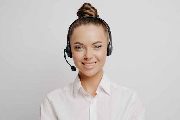 Glimlachende deskundige vrouwelijke callcentermedewerker in wit overhemd met haar in knot en zwarte hoofdtelefoon die graag klanten van dienst is en helpt, alleen voor grijze achtergrond