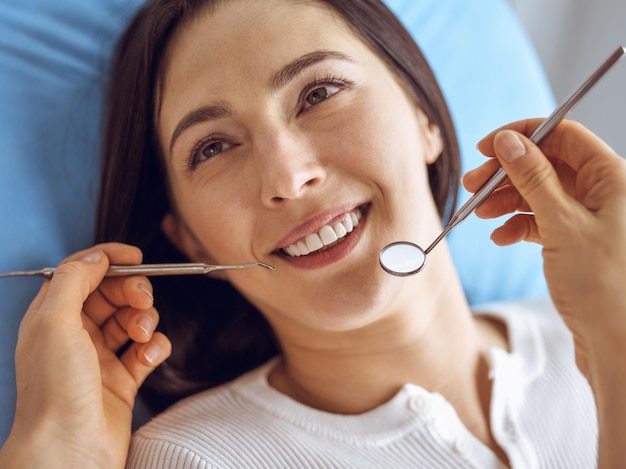 Glimlachende donkerbruine vrouw die door tandarts bij tandkliniek wordt onderzocht. Handen van een arts die tandheelkundige instrumenten vasthoudt in de buurt van de mond van de patiënt. Gezonde tanden en geneeskunde concept.