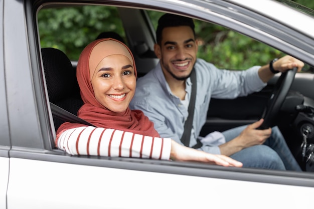 Glimlachende duizendjarige man aan het stuur en vrouw in hijab rijden in de auto genieten van samen reizen