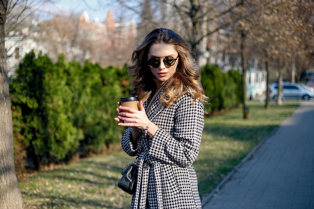 Glimlachende elegante vrouw in trenchcoat wandelen en koffie drinken uit papieren beker. lachende vrouw met krullend haar kijk naar camera glimlach wandelen slow motion gezicht zonsondergang mooie dame buiten close-up schattig