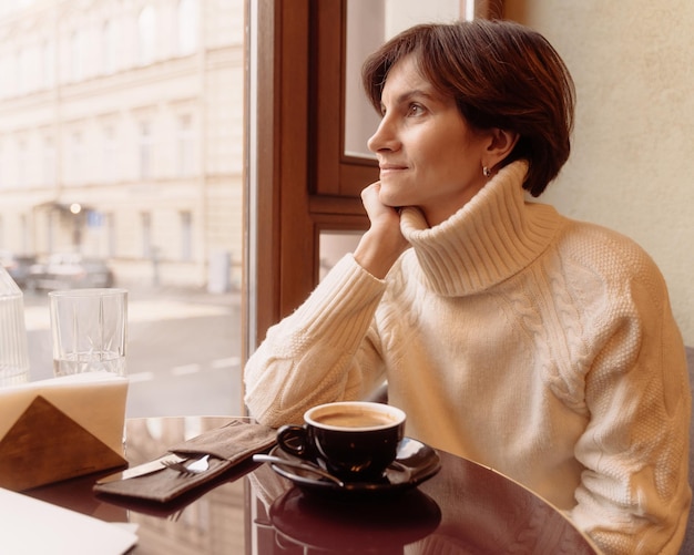 Glimlachende en bedachtzame vrouw in trui die naar het raam in café kijkt terwijl ze koffie drinkt
