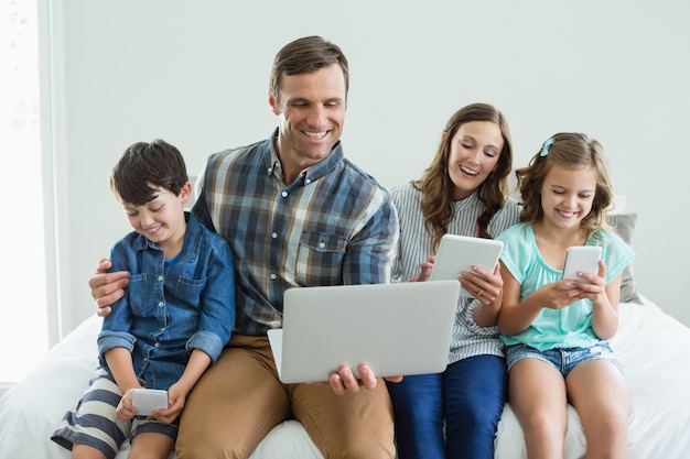 Glimlachende familie met laptop, digitale tablet en mobiele telefoon in de slaapkamer