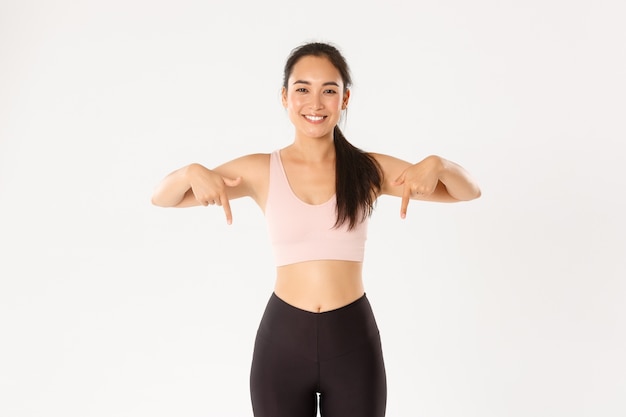 Glimlachende fit en slank vrouwelijke fitnessinstructeur, Aziatisch meisje trainen in de sportschool en iets tonen, vingers naar beneden, staande witte achtergrond.