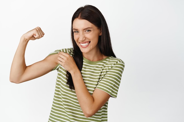 Glimlachende fitte en gezonde vrouw die biceps buigt en sterke arm toont met spier die tegen een witte achtergrond staat in gestreepte tshirt