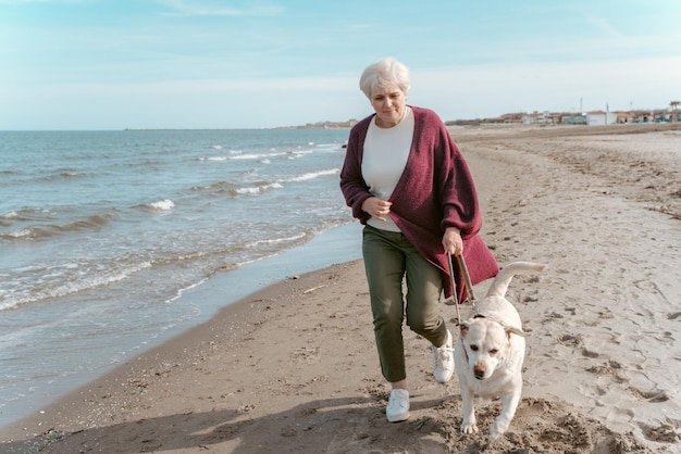 Glimlachende, gelukkige, aantrekkelijke oudere vrouw die met haar schattige Labrador Retriever langs het zandstrand wandelt