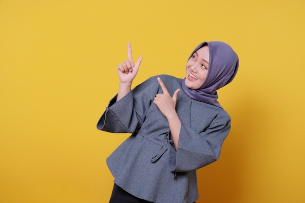 Glimlachende gelukkige Aziatische vrouw die hijab draagt met haar vinger wijzend geïsoleerd op lichtgele bannerachtergrond
