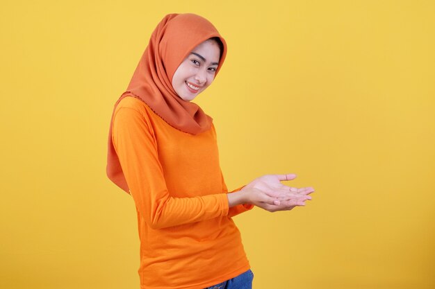 Glimlachende gelukkige aziatische vrouw iets in haar hand en presenterend op lichtgele bannerachtergrond die hijab draagt