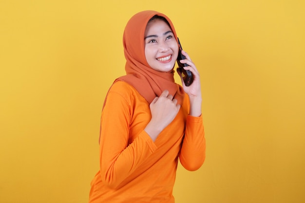 Glimlachende gelukkige aziatische vrouw met geïsoleerde op lichtgele bannerachtergrond die hijab draagt, die met mobiele telefoon spreekt