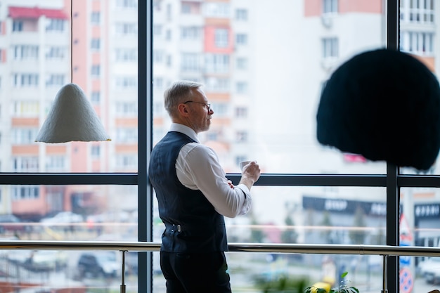 Glimlachende gelukkige directeur denkt aan zijn succesvolle loopbaanontwikkeling terwijl hij met een kopje koffie in zijn hand in zijn kantoor in de buurt van de achtergrond van een raam met kopieerruimte staat