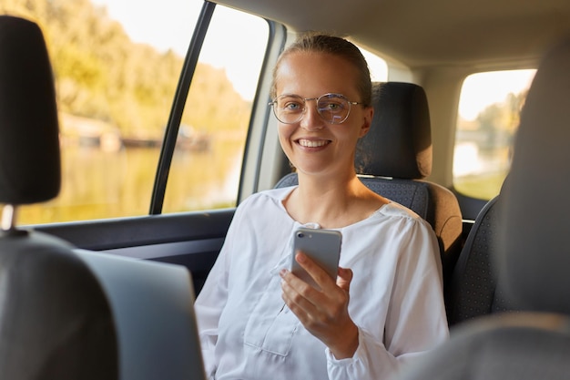 Glimlachende gelukkige positieve vrouw gebruikt een slimme telefoon en glimlacht terwijl ze op de achterbank in de auto zit en mobiele telefoon gebruikt terwijl ze naar de camera kijkt voor zakenreizen