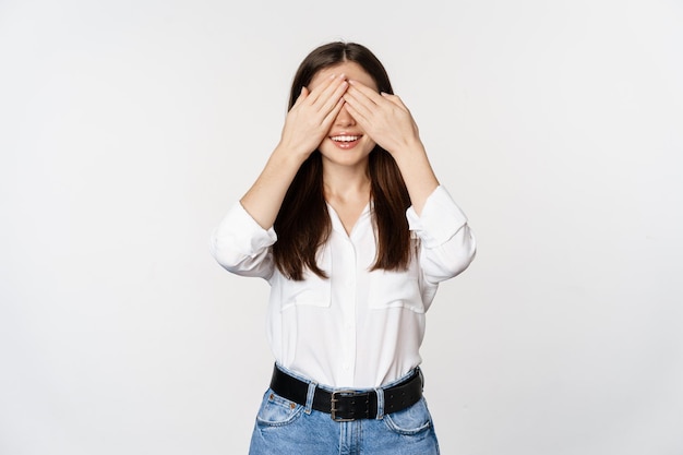 Glimlachende gelukkige vrouw die op verrassing wacht, ogen met handen sluit, blindelings tegen witte achtergrond staat