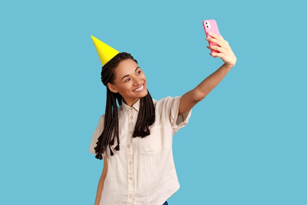 Glimlachende gelukkige vrouw in partijkegel die livestream-opnamevideo uitzendt voor blog tijdens de viering van haar verjaardag met een wit overhemd Indoor studio-opname geïsoleerd op blauwe achtergrond