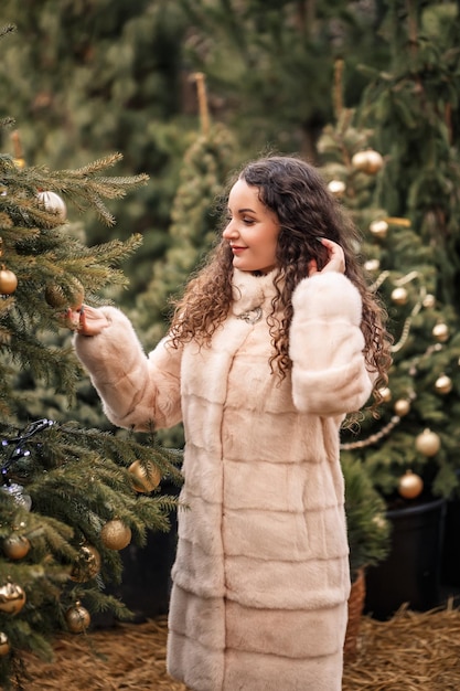 Glimlachende gelukkige vrouw met krullend haar in een bontjas wandelt door de kerstboommarkt wachtend op kerstmis en nieuwjaar