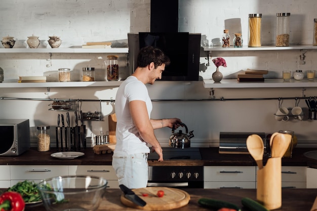Glimlachende gespierde man in wit t-shirt die waterkoker op de oven in de keuken zet