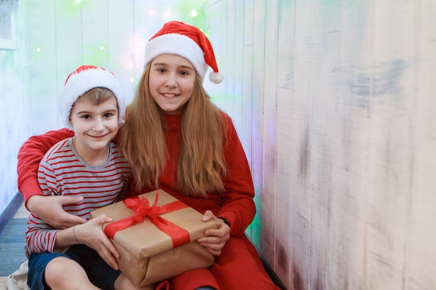 Glimlachende grappige twee kinderen die in de rode kerstmuts van de Kerstman de gift van Kerstmis in hand houden. Nieuwjaar concept.