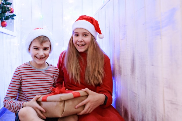 Glimlachende grappige twee kinderen die in de rode kerstmuts van de Kerstman de gift van Kerstmis in hand houden.