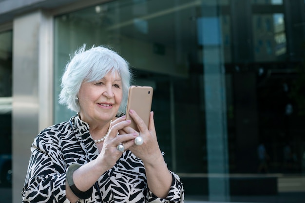 Glimlachende grijsharige oudere vrouw met mobiele telefoon in haar hand op straat