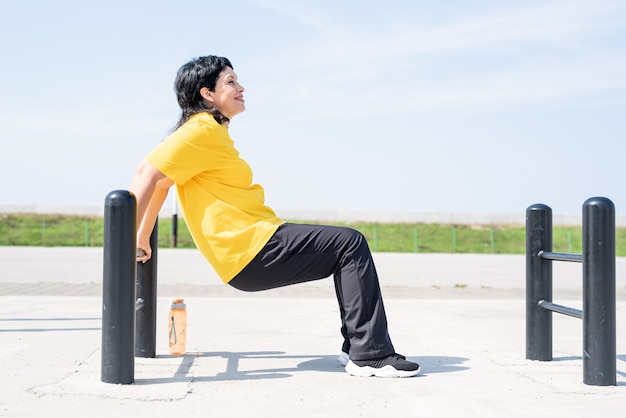 Glimlachende hogere vrouw die omgekeerde push-ups buiten op de sportveldbalken doet