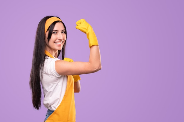 Glimlachende huisvrouw in schort en handschoenen die bicepsen tonen