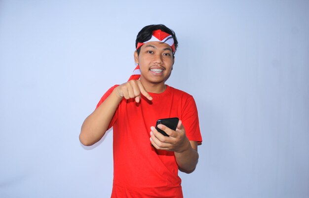Glimlachende indonesische jongeman die naar mobiele telefoon wijst met een blij en opgewonden gezicht in een rode t-shirt