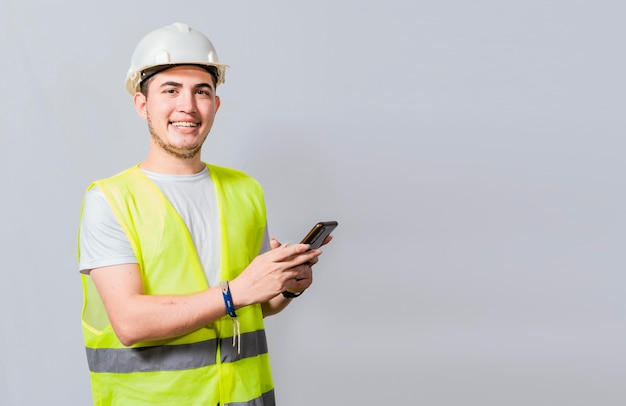 Glimlachende ingenieur die mobiele telefoon gebruikt die camera bekijkt Vriendelijke mannelijke bouwer-ingenieur die telefoon gebruikt en camera bekijkt Jonge ingenieur in vest en helm die mobiele telefoon gebruikt die camera glimlacht