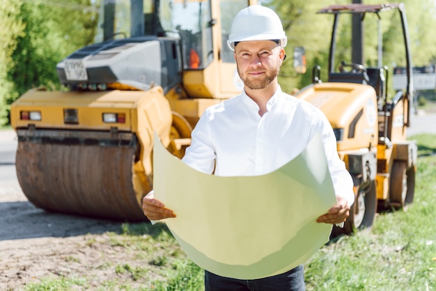 Glimlachende ingenieur met helm die zich voor graafwerktuig op de bouwplaats van de weg bevindt