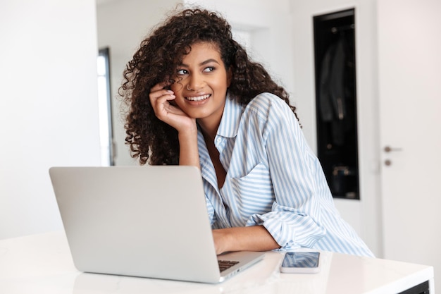 glimlachende jonge afrikaanse vrouw die op een laptop aan de tafel thuis werkt, met behulp van mobiele telefoon