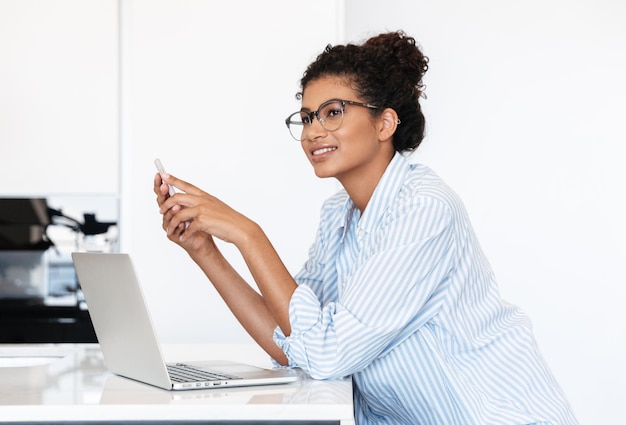 glimlachende jonge afrikaanse vrouw die op een laptop aan de tafel thuis werkt, met behulp van mobiele telefoon