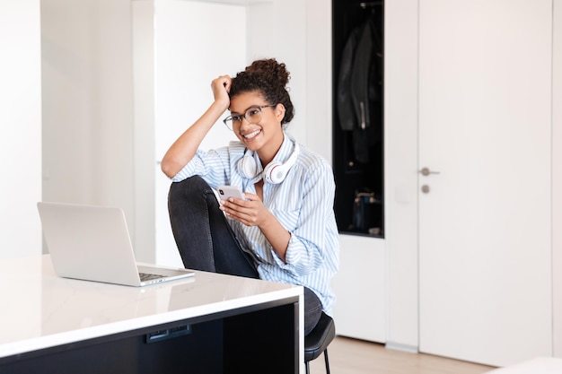 Glimlachende jonge afrikaanse vrouw die op een laptop aan de tafel thuis werkt, met behulp van mobiele telefoon