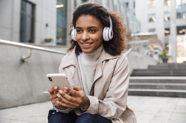 Glimlachende jonge afrikaanse vrouw met een herfstjas die op een trap op straat zit en een mobiele telefoon gebruikt terwijl ze naar muziek luistert met een draadloze koptelefoon