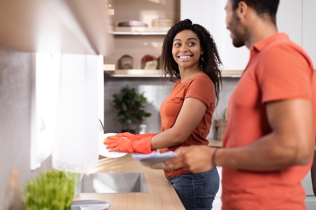 Glimlachende jonge Afro-Amerikaanse man en vrouw in dezelfde t-shirts die samen borden wassen in het keukeninterieur