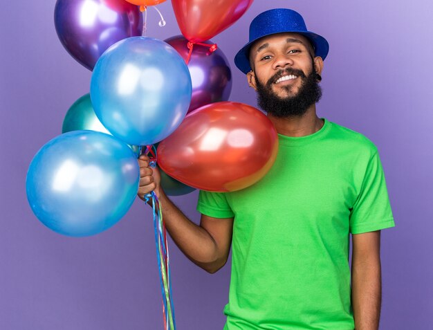 Glimlachende jonge Afro-Amerikaanse man met een feestmuts met ballonnen geïsoleerd op een blauwe muur