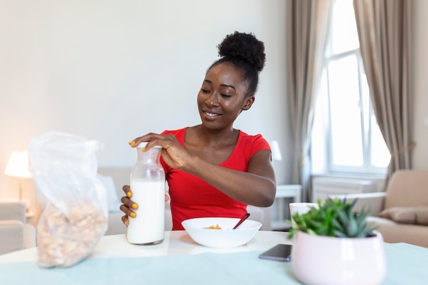 Glimlachende jonge Afro-Amerikaanse vrouw giet cornflakes in bord met melk Het meisje heeft 's ochtends een gezond ontbijt op een stijlvol, gezellig huis terwijl ze haar e-mail controleert op laptop