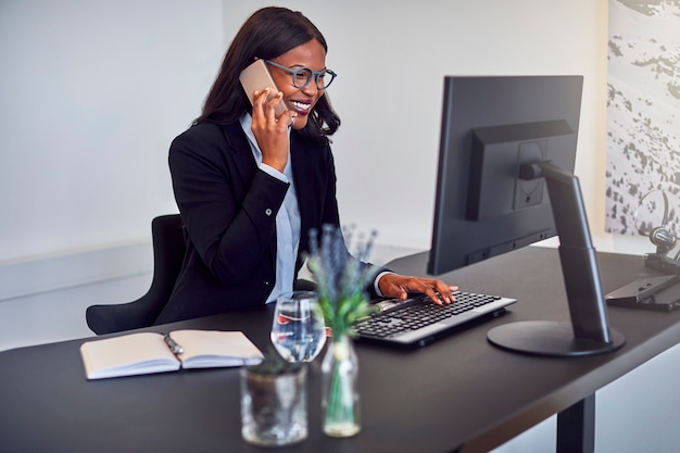 Glimlachende jonge Afro-Amerikaanse zakenvrouw die op een mobiele telefoon praat terwijl ze op een computer werkt