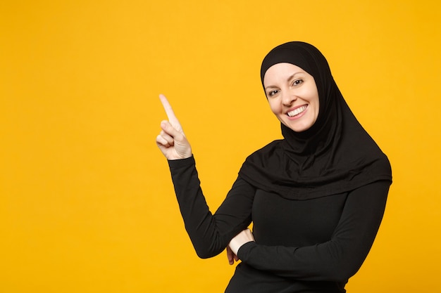 Glimlachende jonge Arabische moslimvrouw in hijab zwarte kleding met wijzende kopie ruimte met handen vinger geïsoleerd op gele muur portret. Mensen religieuze levensstijl concept.