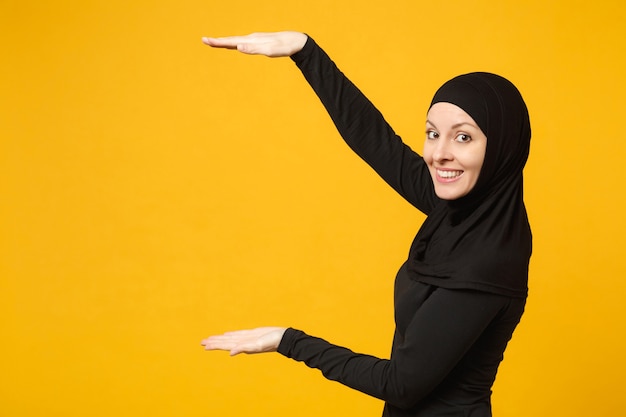 Glimlachende jonge Arabische moslimvrouw in hijab zwarte kleding met wijzende kopie ruimte met handen vinger geïsoleerd op gele muur portret. Mensen religieuze levensstijl concept.