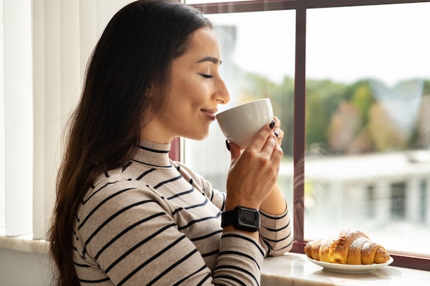 Glimlachende jonge Arabische vrouw drinkt een kopje warme koffie thee met croissant in de lichte woonkamer