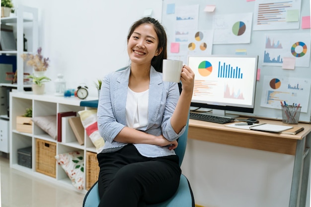 glimlachende jonge Aziatische Chinese vrouwelijke ondernemer die een kopje koffie drinkt en op de computer werkt terwijl hij aan het bureau zit in een helder thuiskantoor. elegant meisje freelance werknemer gezichtscamera geniet van theepauze