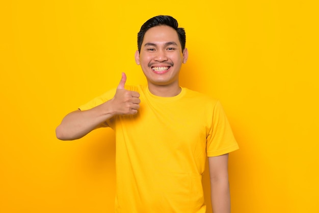 Glimlachende jonge Aziatische man in casual tshirt duim opdagen en gebaren teken van goedkeuring geïsoleerd op gele achtergrond Mensen lifestyle concept