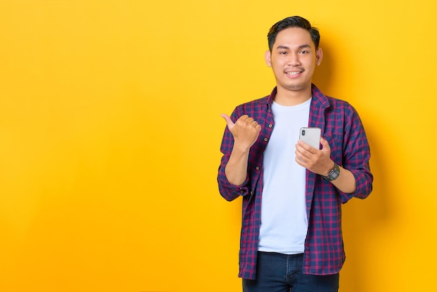 Glimlachende jonge Aziatische man in geruit hemd met bankbiljetten en wijzende vingers naar kopieerruimte geïsoleerd op gele achtergrond