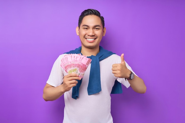 Glimlachende jonge Aziatische man met witte t-shirt die contant geld in rupiah bankbiljetten vasthoudt en duimen omhoog gebaart geïsoleerd over paarse achtergrond mensen levensstijl concept