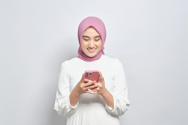 Glimlachende jonge Aziatische Moslimvrouw die mobiele telefoon gebruikt die goed nieuws krijgt dat op witte achtergrond wordt geïsoleerd