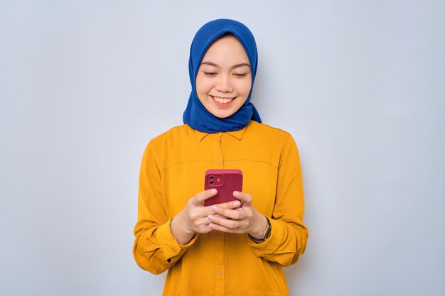 Glimlachende jonge Aziatische moslimvrouw gekleed in oranje shirt met behulp van mobiele telefoon typen van SMS-berichten geïsoleerd op witte achtergrond