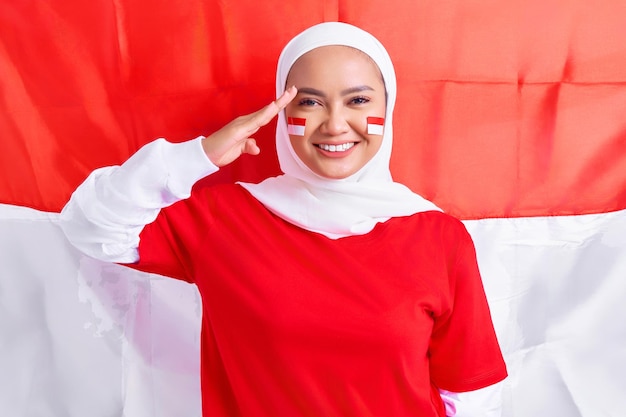 Glimlachende jonge Aziatische moslimvrouw in rood wit tshirt vieren Indonesische onafhankelijkheidsdag op 17 augustus en tonen respect gebaar geïsoleerd op vlag Indonesische achtergrond