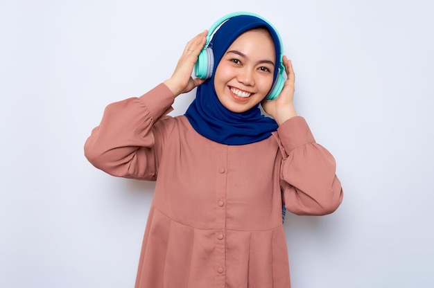Glimlachende jonge Aziatische moslimvrouw in roze shirt luisteren muziek in koptelefoon kijken camera geïsoleerd op witte achtergrond Mensen religieuze levensstijl concept