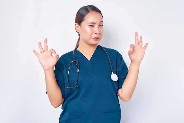 Glimlachende jonge Aziatische verpleegster die een blauw uniform draagt met een stethoscoop verzekert de kwaliteit van de testresultaten in hun klinieklaboratorium met een goed teken geïsoleerd op een witte achtergrond
