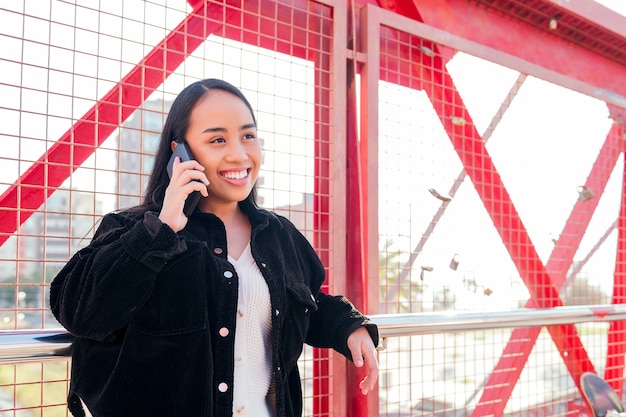 Glimlachende jonge aziatische vrouw die in openlucht telefoneert en tegen een rood hek leunt