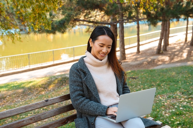 Glimlachende jonge Aziatische vrouw die jas draagt, zittend op een bankje in het park, die op laptop computer werkt